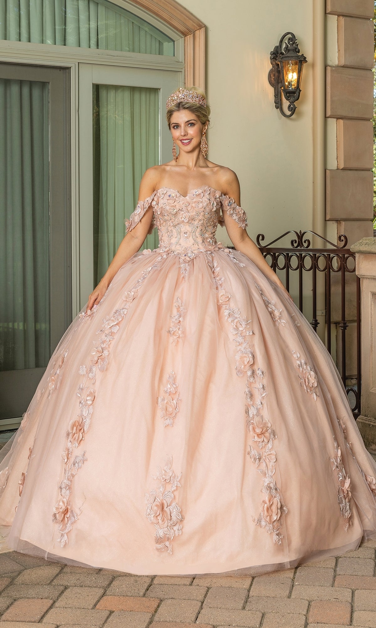 A young woman smiles at the camera outside, wearing Quinceanera Dress 1687 By Dancing Queen in rose gold with floral appliqué, off-the-shoulder sleeves, and a tiara.